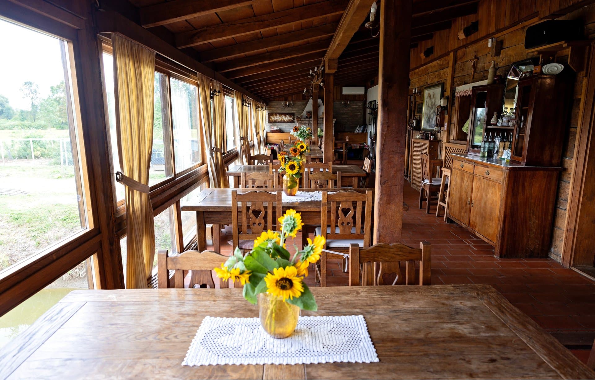 Rustikalni interijer restorana — drveni stolovi, sunčanice i pogled na zelenilo